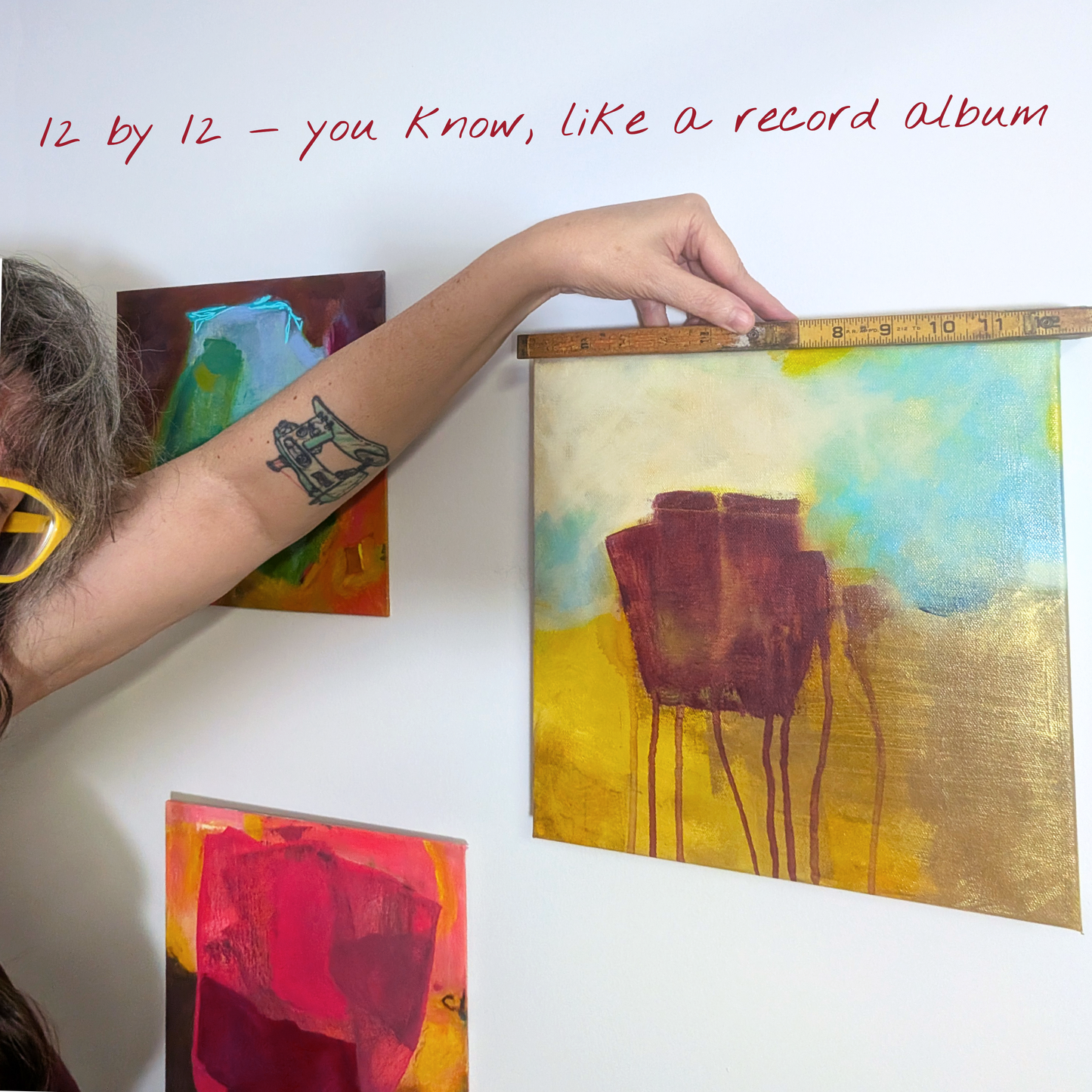 A person's arm is shown holding a wooden ruler against a canvas painting that features yellow, gold, and blue abstract colors. The text '12 by 12 - you know, like a record album' is written in red at the top, and other colorful paintings are visible on the white wall behind.