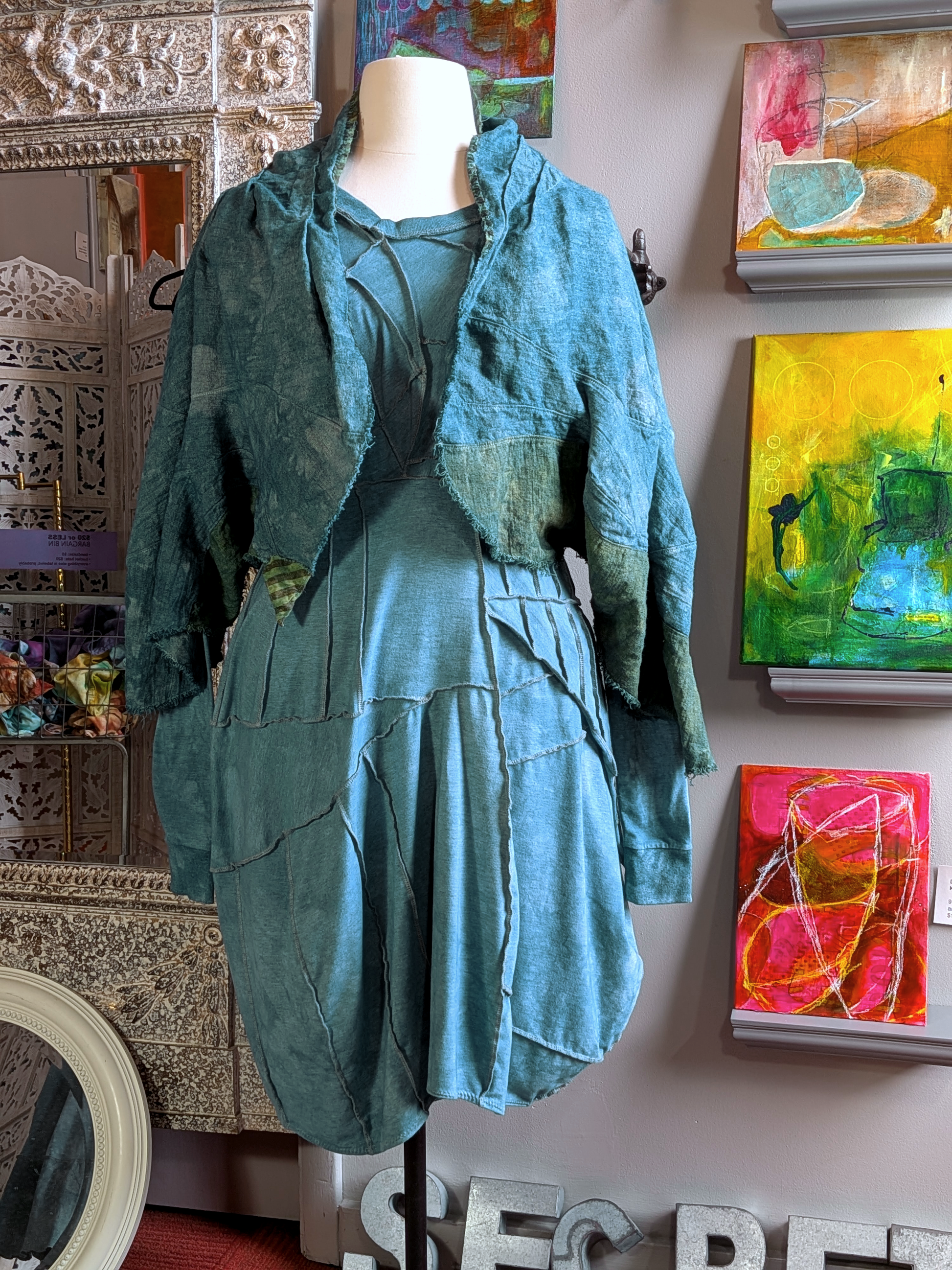 Mannequin wearing a teal dress with a matching jacket in front of colorful abstract paintings. secret lentil
