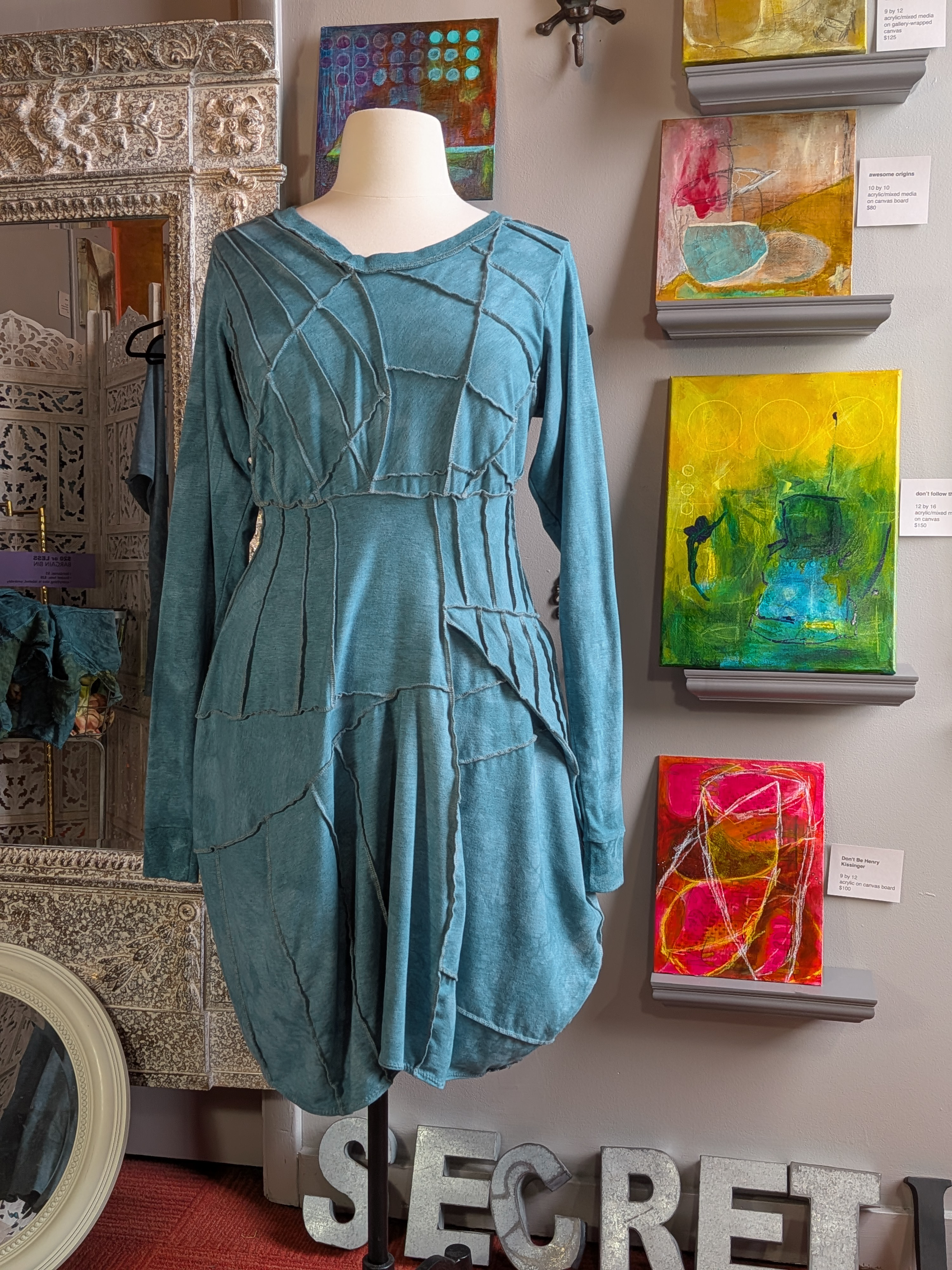 Teal dress on a mannequin in front of wall art with 'Secret Lentil' branding.